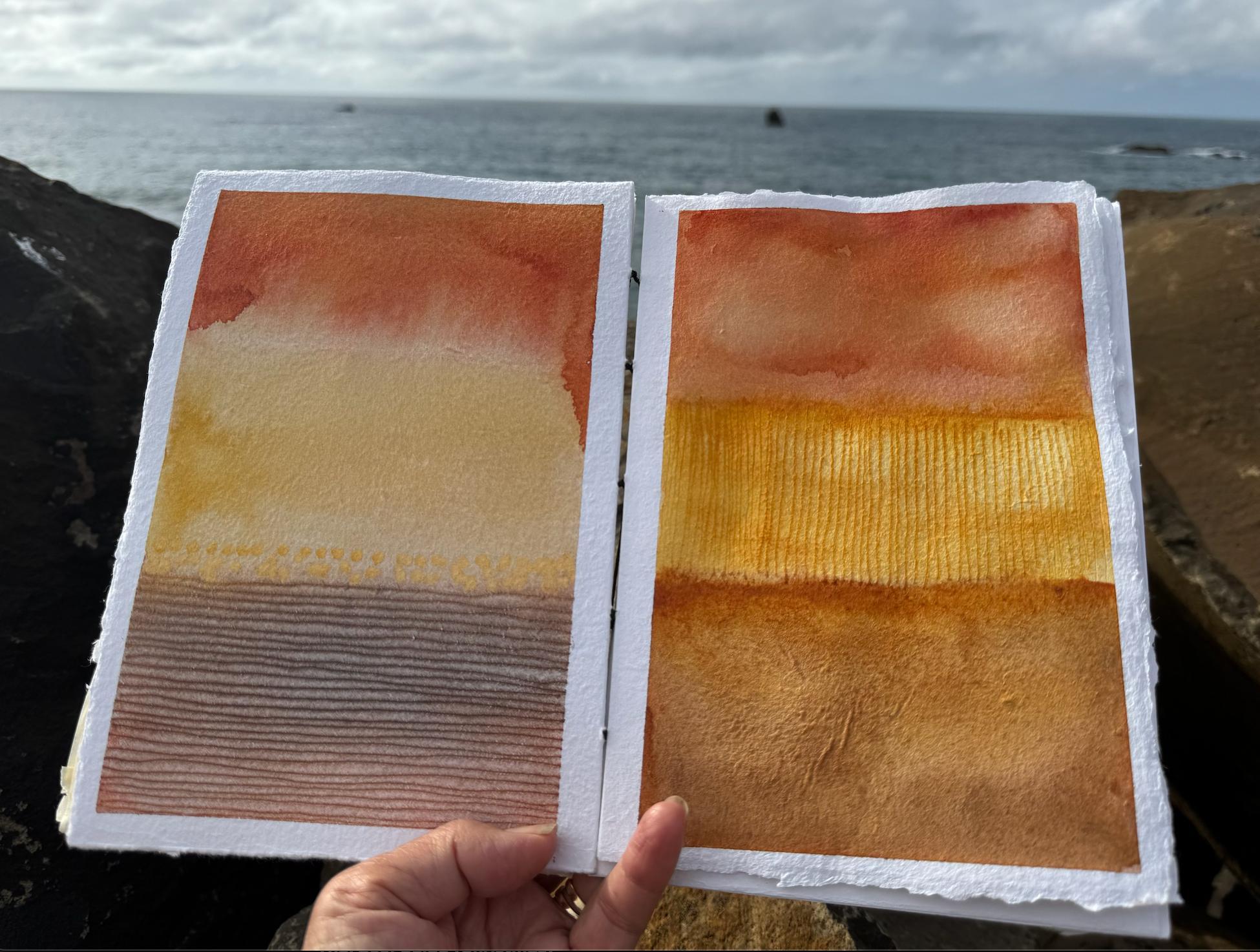 Open art journal held against the ocean, warm terracotta and gold watercolour washes on the pages