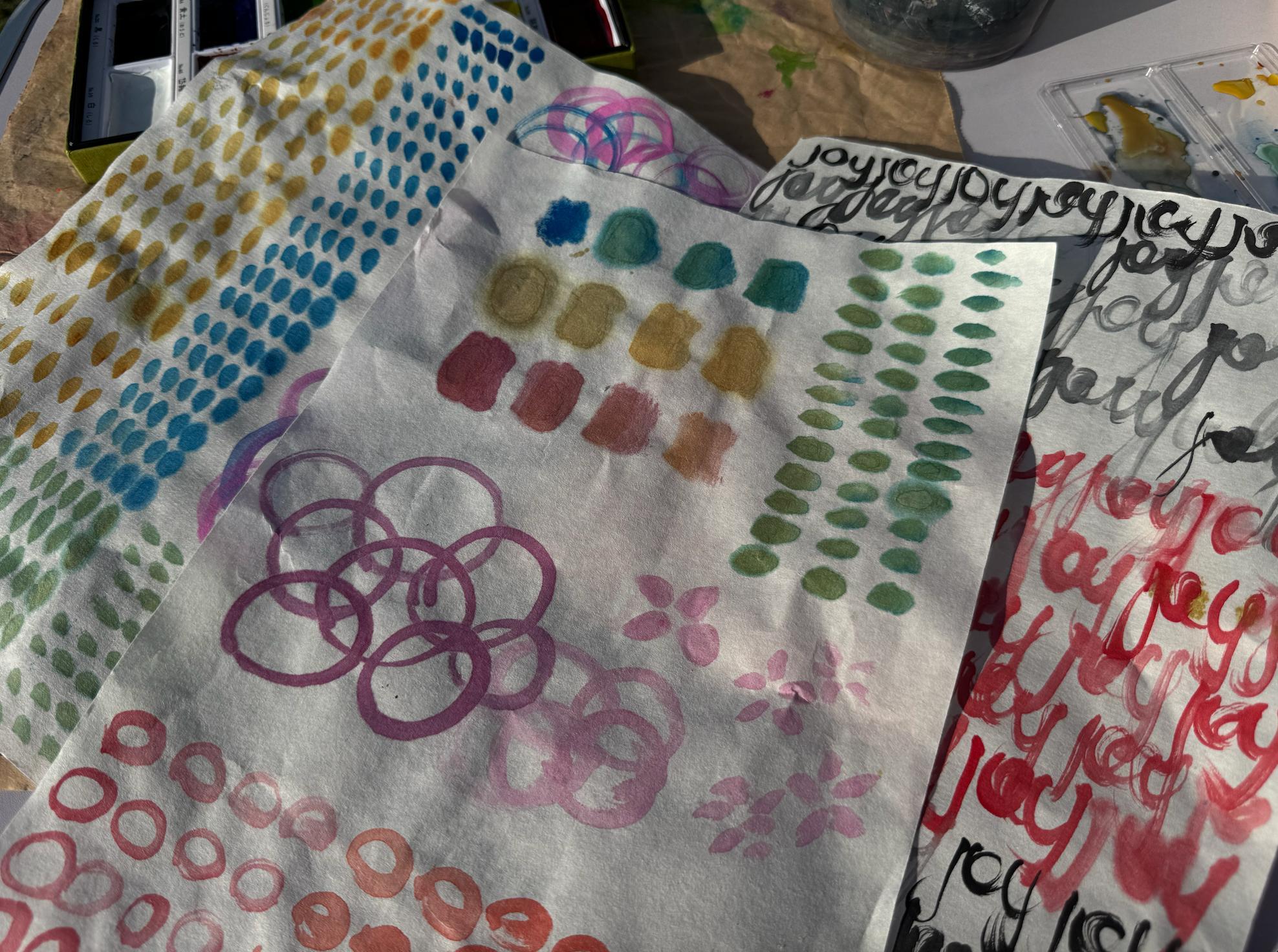 Colourful art journal practice pages laid out in the sun with rainbow dots, circles and joy written in flowing script