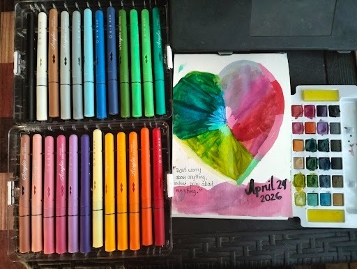 Flat lay of colourful Staedtler marker sets and a watercolour palette alongside an open art journal showing a painted heart in greens, yellows and pinks dated April 29 2026