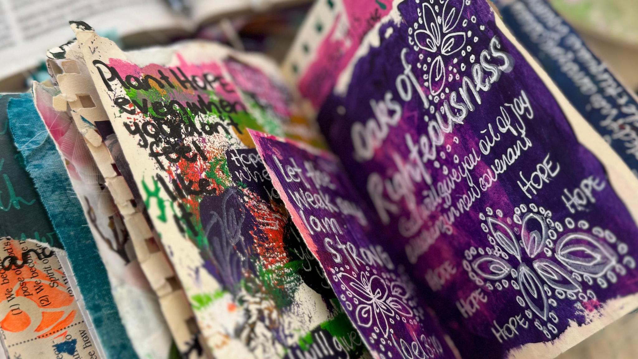 Tina's art journal pages fanned out, showing colourful handwritten words and paint marks